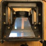 The mirror for the viewing lens/ground glass. The mirror is mounted with clips and glue to a backing plate. The entire backing plate can be removed from the camera. The pairs of nuts at the top and bottom of the photo are the camera strap hangers.