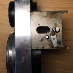 Right side of the lens panel assembly. The rod connecting the focusing wheels goes through the oval holes. The partial pin and lever on the right engage the cam in the focusing knob and move the panel in and out as the knob is turned.